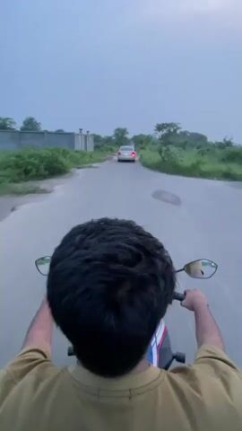 The video presents a first-person view from behind a person riding a motorcycle on a narrow, worn asphalt road. The rider, seen from the head and shoulders, is wearing a light brown shirt and has dark, curly hair. Their hands are on the handlebars, with the motorcycle's rearview mirrors visible on either side.

Ahead on the road, a silver sedan is being followed. The road surface shows signs of wear and contains several puddles, indicating recent rain. Lush green trees and bushes line both sides of the road, and a concrete wall is visible on the left. The sky is overcast, suggesting it is either dusk or a heavily cloudy day. The scene depicts a journey through a rural or semi-rural landscape, consistent with Purbachal, Bangladesh. No visible text is present.