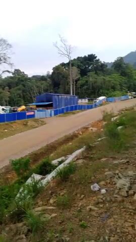 The video shows a construction site in a hilly, wooded area. In the foreground, there is a dirt road and some overgrown vegetation. Behind the road, there is a blue construction fence with machinery and vehicles visible beyond it. Further in the distance, there are power lines and a transmission tower against a backdrop of lush green mountains and a cloudy sky. The video pans across the landscape, offering views of the construction site and the surrounding natural environment. There are no people visible in the video.