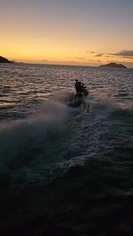 A wide shot depicts a person riding a jet ski on the ocean at sunset. The sun is setting, casting a golden glow on the horizon. In the background, there is a dark land mass that resembles an island. The jet ski is moving through the water, creating white waves and a trail of foam. The ocean water is dark and choppy. A person can be seen riding the jet ski, but their features are not distinct. A musical rhythm can be heard in the audio.