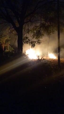 At night, two piles of debris burn, creating a large amount of smoke. The flames cast light over the dry ground and illuminate nearby trees. A wooden structure, possibly a frame, lies near one of the fires. A post stands between the viewer and the fires.