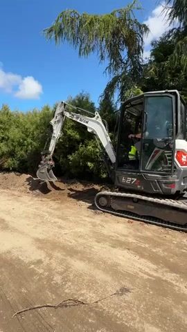 A white and gray Bobcat E27Z mini-excavator is operating outdoors on a dirt surface. The excavator has a black operator's cab with a person inside wearing a high-visibility vest. The excavator's boom is extended and digging into a pile of dirt and vegetation on the left side of the frame. Lush green trees and foliage are visible in the background. The sky is blue with scattered white clouds. A long, thin, brown object, possibly a root or branch, is lying on the dirt in the foreground. The video is short and shows the excavator in motion.
