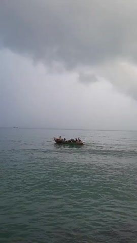 A small boat with four people is moving across the ocean on a cloudy day. The boat is traveling away from the viewer. The water is dark. In the far distance, there is a ship on the horizon. The sky is overcast with dark gray clouds. The edge of the frame shows a few small waves near what may be a rocky shoreline.