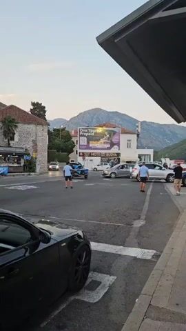 A man in a blue shirt and shorts is standing in a street intersection, looking at a dark blue SUV that is stopped in the road. The SUV has its brake lights on. There are other cars and a billboard in the background. The sky is a light gray, and there are mountains in the distance. The time is approximately evening.
