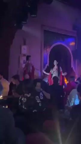 The video captures a vibrant nightclub atmosphere.  On a raised platform, two dancers, one wearing a red shawl, perform for a crowd of people. The club is illuminated by blue and orange lighting, accentuated by a large chandelier.  In the background, a band can be seen playing. Some people in the crowd are standing while others sit at tables, where bottles are visible.