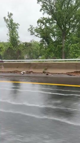 The video depicts a yellow Chevrolet Corvette driving on a wet highway. The car has black rims and a black roof. Raindrops cover the car's surface. Other vehicles, including a white sedan and a semi-truck, can be seen in the background on the opposite side of the highway. The background features green trees and an overcast sky. The road has yellow lane markings.