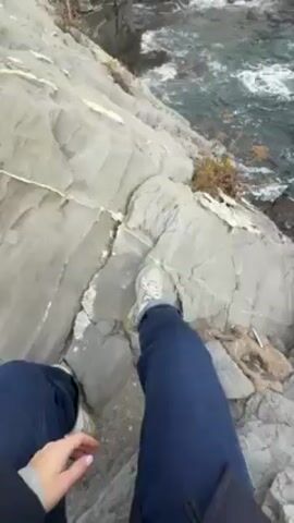 A first-person view shows a person sitting on a rocky cliff overlooking the sea. The person is wearing blue jeans and white sneakers. The camera pans up to reveal the ocean with waves crashing against the rocks. In the background are buildings and trees along the coast. The sky is cloudy.
