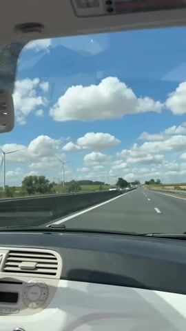 The video begins with a view from inside a car, looking out through the windshield. The road ahead is a highway with multiple lanes and other vehicles. The sky is blue with fluffy white clouds, and in the distance, wind turbines are visible. The car's dashboard and part of the steering wheel are visible in the foreground. The perspective then shifts to a side view from inside the car, showing a green guardrail alongside the road. Finally, the video shows a close-up of a woman with blonde hair wearing sunglasses and a pink top. She is smiling and has her arm raised in the air, with her hair blowing in the wind, suggesting she is enjoying a ride in a convertible or with the windows down.