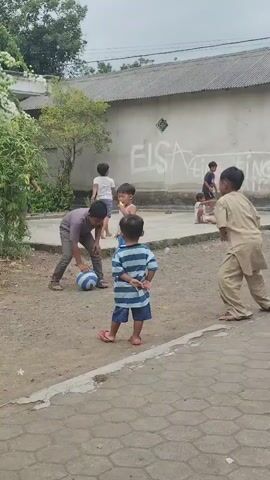 Several young boys are playing soccer in a dusty outdoor area. The boy in the foreground, wearing a blue and white striped shirt and blue shorts, stands facing away from the camera with his hands behind his back. To his left, a boy in a grey shirt and pants kicks a blue and white soccer ball. Other boys are scattered around the area, some watching and some actively participating in the game. One boy in a tan outfit appears to be anticipating a pass. In the background, a white wall is marked with graffiti, including the name "ELSA." The area is unpaved, with some vegetation to the left and a tiled walkway in the foreground. The sky is overcast.