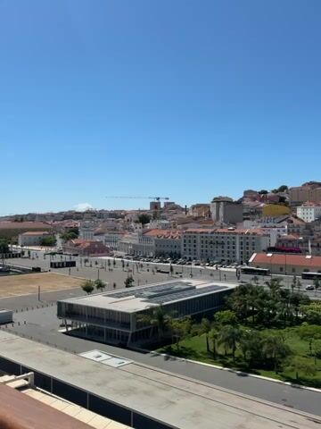 This is a vertical video clip offering a panoramic view of Lisbon, Portugal, under a clear blue sky. The scene captures a bustling cityscape dominated by buildings with terracotta roofs, characteristic of Lisbon's architecture, cascading down a hill.

In the foreground, a modern, rectangular building with a glass facade is situated amidst a manicured green space with palm trees. Adjacent to this, a large, flat, concrete plaza stretches across the frame, suggesting a public space or a wide walkway. A body of water, possibly a reflecting pool or a decorative pond, is visible to the right of the modern building, reflecting the surrounding architecture and the sky.

Further back, the dense urban fabric of Lisbon unfolds. Numerous buildings, varying in height and color, fill the midground and background, creating a picturesque urban landscape. A prominent dome and spire, likely belonging to a church or cathedral, rise above the rooftops in the distance, a key landmark of the city. Construction cranes are also visible on the horizon, indicating ongoing development.

The time of day appears to be daytime, with bright sunlight casting distinct shadows. The clear sky suggests fair weather. While the video is a wide shot, there are hints of activity: cars and buses can be seen on the streets, and small figures appear to be walking in the plaza and along the sidewalks, indicating a normal day in the city. There is no visible text in the video. The overall emotion evoked is one of warmth, vibrancy, and the historical charm of a European capital city.