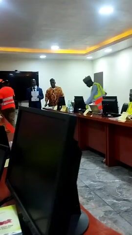 Here is a detailed description of the video:

The video appears to be taken inside an office or meeting room. There are multiple people present, some wearing reflective vests, suggesting they may be related to construction or field work.  The room is well-lit, with what seems to be recessed lighting in the ceiling. There is a large screen on the wall at the far end of the room. A large table stretches across the room, with computer monitors, laptops, water bottles, and Coca-Cola bottles placed on it. The individuals are either seated at the table or standing around it, seemingly engaged in a meeting or training session. There is a man in a brown patterned shirt standing and talking at the head of the table, and someone else in a suit standing beside him holding a piece of paper. Some of the individuals at the table are reviewing documents. The focus of the room is on the work at hand with everyone engaged.