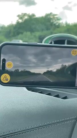 The video provides a first-person perspective from inside a car driving on a road. A black smartphone is mounted horizontally on the vehicle's black, textured dashboard, which features white stitching.

The phone's screen displays a live view of the road ahead, mirroring the scene visible through the car's windshield. On the phone screen, several circular icons are present: a green dot accompanied by the number "277" in the top left, a yellow triangle with an exclamation mark and a plus sign in the bottom left, and a yellow map icon in the top right.

The view through the windshield shows a two-lane asphalt road extending into the distance. The road is marked with a solid white line on the right shoulder and a broken white line in the center. Lush green trees and dense vegetation flank both sides of the road. Several vehicles are visible in the distance on the road.

The sky is predominantly cloudy, with various shades of grey clouds and some brighter patches, including fluffy white cumulus clouds on the horizon. The lighting suggests it is daytime. The setting appears to be a rural or semi-rural area, consistent with the provided context of Lugoj, Romania. Background music is playing throughout the video.