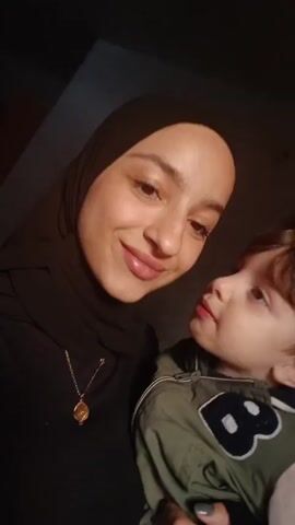 A woman wearing a black hijab holds a young child, likely her son. The child, with light brown hair, has his face partially turned towards the camera. The mother and child are close, with the child nuzzling his face against the woman's cheek. As the music plays, the woman angles the camera for a closer view of their faces. The child looks sleepy and yawns, and the woman makes a kissing face at the camera.