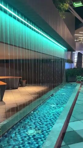 A long, narrow water feature with blue tiled bottom and a curtain of water flowing down from above, illuminated by green LED lights. The water feature is bordered by a tiled walkway. In the background, there is a modern building with multiple levels, shops, and signage, including a large "JUST DO IT" sign. A few plants are visible. The lighting suggests it is evening or night.