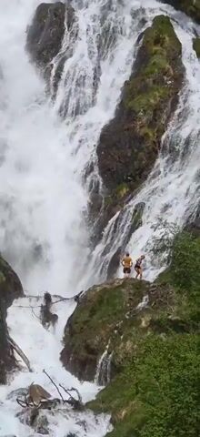 The video features a large, multi-tiered waterfall cascading down a mountain covered with lush green vegetation. Two people can be seen standing on a rocky outcrop near the base of the waterfall. The camera zooms out, revealing a wider view of the waterfall and its surroundings, including a bicycle parked in the foreground. The sky is visible in the background, with scattered clouds. The overall environment appears to be a natural, scenic mountain area.