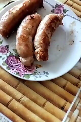 The video shows a close-up of cooked sausages on a plate. Then it pans to a bowl of chopped salad, which appears to be cucumber, onion, and dill in a creamy dressing. Finally, it shows a bowl of yellow cherries. The background suggests a casual setting, possibly a kitchen or dining area, with a bamboo mat visible.