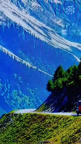 A video shows a mountainous landscape with a winding road and a vehicle. The mountains are covered in snow and blue hues. The road is paved and curves through lush green grass. There are pine trees on the right side of the road, and a dark-colored vehicle, possibly a truck or bus, is parked on the road. The scene appears to be an outdoor scenic view.