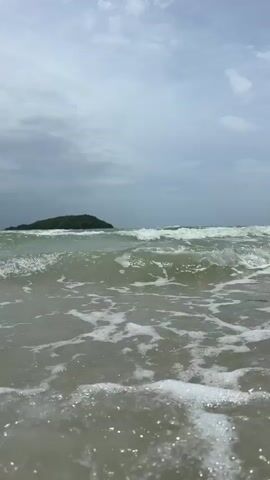 A low-angle shot shows ocean waves rolling onto a sandy beach. In the distance, a small, tree-covered island is visible under a cloudy sky. The water is a muted green-grey color with white foam from the breaking waves. The camera is positioned just above the water's surface, capturing the movement of the waves and the foam washing ashore.
