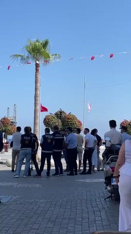 A group of people are gathered outdoors on a sunny day. Several individuals appear to be police officers, identifiable by their vests with the word "POLİS" printed on them. One officer in the foreground faces away from the camera, while others in the middle ground are in conversation. Behind them, a tall palm tree stands prominently against a clear blue sky, adorned with red and white triangular flags strung between poles. A Turkish flag is visible on the palm tree, and another flag, possibly the flag of Northern Cyprus, flies from a flagpole to the right. Other individuals, seemingly civilians, stand in smaller groups or individually, observing the scene. The ground is paved with light-colored cobblestones. In the background, there are glimpses of structures and possibly a body of water. The overall impression is of a public gathering or event taking place in a coastal or waterfront area.