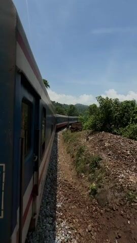 A train car is seen moving along a track that curves out of view. The car is blue with white and red stripes. Outside the car, the track is lined with gravel. Beyond the gravel, there's a small dirt embankment with some sparse vegetation and piles of rusted metal. In the background, there's dense tropical vegetation under a partly cloudy blue sky.