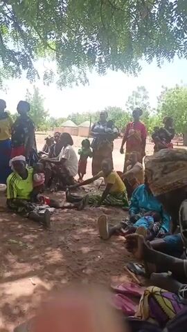The video appears to show a gathering of people in an outdoor, rural setting, possibly a village. There are men, women, and children present.  Some individuals are sitting on the ground, while others are standing. Some women are carrying babies.  A few men are seated on plastic chairs.  A man with a beard, wearing sunglasses and a cap, is standing and appears to be addressing the group.  There are trees providing shade. The ground is dirt.  In the background, there are simple buildings or huts. A red vehicle is also visible in the distance.  The general atmosphere seems relaxed and informal.