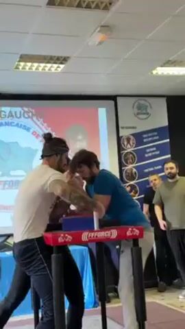 Here's a description of the video:

Two men are arm wrestling. One man has his hair in a bun with a bandana, is wearing a white t-shirt, and has tattoos on his arm. The other man has dark curly hair and is wearing a blue t-shirt. They are at a red arm wrestling table with the brand name "FFFORCE" on it. Behind them is a projection screen with a logo on it. There are a few other people watching in the background. The focus is on the arm wrestling match, with the camera zooming in slightly as the competition starts.
