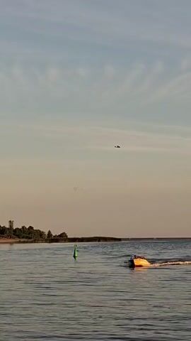 A drone flies high in the sky over a calm body of water. In the foreground, a small yellow boat with white trim is speeding across the water, leaving a wake behind it. The boat appears to be in motion, possibly a jet ski or a small speed boat. There is a green buoy in the water to the left of the frame. The sky is a light blue with wispy white clouds. The horizon is visible in the distance, separating the water from the sky. The overall scene suggests a serene day on the water, with the drone adding a modern element to the natural landscape.