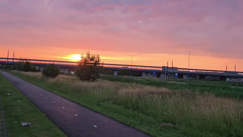 The video shows a sunset over a rural landscape with a bicycle path in the foreground. The sky is filled with orange and pink clouds as the sun sets behind a highway overpass in the distance. Tall grass and a few trees are visible in the midground. The overpass has several lights on, and some vehicles are visible on the road. The bicycle path is paved and has white lines marking the lanes. The video is shot from a slightly elevated angle, looking towards the sunset.