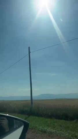 This media file is a vertical, point-of-view video recording taken from inside a vehicle. The scene is a bright, sunny daytime view of a rural landscape. 

**Subjects:**
*   **Objects:** The primary objects visible are the side mirror of the vehicle in the foreground, a power pole with a single wire stretching across the frame, and a distant mountain range. The vehicle's interior, including a hand holding a phone, is reflected in the side mirror.
*   **Scene/Setting:** The setting is a wide-open rural area, likely Târgu Secuiesc, Romania. It features a field of dry, tall grass or crops adjacent to a grassy roadside. The sky is a clear, vibrant blue, indicating good weather. In the distance, a range of mountains forms the horizon, and the sun is high and bright, casting lens flare and illuminating the scene.

**Activity or Event:**
The video appears to capture a journey or a scenic drive through the countryside. The movement of the vehicle is implied by the perspective.

**Notable Details:**
*   **Time of Day:** Midday, due to the high position of the sun and the intense brightness.
*   **Weather:** Clear and sunny with no visible clouds, suggesting warm weather.
*   **Location Cues:** The rural landscape with fields, a power line, and distant mountains are characteristic of many rural areas, and specifically Târgu Secuiesc, Romania as indicated.
*   **Emotions:** The bright sunlight and expansive view evoke feelings of openness, freedom, and perhaps a sense of travel or vacation.
*   **Visible Text:** There is no visible text in this media file.