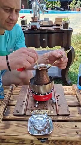 A man in a teal t-shirt is making coffee outdoors. He is seated at a wooden table with a small red portable stove on it. On the stove, a copper pot is heating, and the man is stirring the contents with a spoon. Coffee beans are scattered on the table. In front of the stove, there is a small glass cup on a saucer. In the background, there is a swimming pool and a wooden table with a coffee maker and plants.