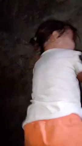 A child, wearing a white t-shirt and orange shorts, is seen in a dimly lit, enclosed space. The child appears to be lying down or in a contorted position, with their back to the camera. The environment is dark and appears to be a rough, textured wall, possibly concrete or stone. The lighting is poor, creating shadows and obscuring details of the surroundings. No other individuals or objects are clearly visible. The activity appears to be rest or sleep, given the child's posture. There are no discernible text elements within the frame. The overall atmosphere is indistinct due to the low light and the limited view of the setting.