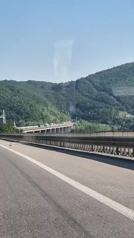 A truck is seen driving on a highway with a large bridge in the background. The bridge spans a valley with forested hills on either side. The sky is clear and blue.