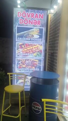 This is a brief video of a food stall at night. The stall has a large illuminated sign that advertises "DÖVRAN DÖNER" and displays pictures of various dishes, including shawarma, koobideh kabob, and mixed kabob. In front of the sign, there is a blue barrel with a Pepsi logo on it, which appears to be used as a table. Two yellow bar stools are placed near the barrel. The background shows a brick wall and some greenery. The lighting suggests it is evening or nighttime, and the overall atmosphere is that of an outdoor fast-food establishment.