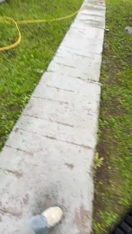 The video is a first-person perspective shot from someone walking. The environment appears to be an outdoor setting, possibly a residential area. In the first frame, a tire is visible in the foreground, and a paved walkway with grass on either side is in the background. The second frame shows a blurry view of houses with red roofs, surrounded by trees and grass. The third frame depicts a paved sidewalk running alongside a building and a grassy area with trees. A power pole and a distant tower are also visible. The overall impression is of a cloudy or overcast day.