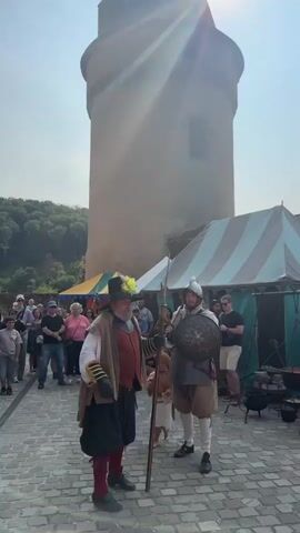 The video shows a historical reenactment taking place in front of a stone tower. Two men in period costumes, one holding a long pole and the other a round shield and a sword, are in the foreground. The man with the pole wears a wide-brimmed hat with a feather, a brown vest over a reddish shirt, and dark breeches. The man with the shield wears a helmet, a chest plate, and breeches. A child in a light-colored tunic and hood is near the man with the pole. A crowd of spectators, dressed in modern clothing, watches the reenactment. Behind the reenactors, there are tents and trees. The ground is paved with cobblestones, and the sky is clear and bright. The sun is shining, casting a lens flare in the upper right corner of the frame.
