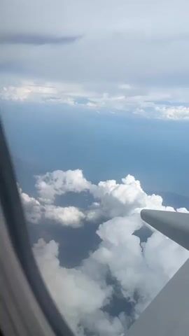 From the window of an airplane, the wing is visible as it passes over a partially cloudy seascape. The wing is grey with multiple sections, ending with a red fin at the back.  Below the plane, white clouds obscure a portion of the landmass.  The sea is a light blue, and the sky is partially covered in grey and white clouds.