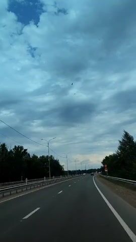 A highway with two lanes in each direction is visible. The road is paved and has white lane markings. On the sides of the highway are guardrails and trees. The sky is cloudy and appears to be a deep blue color. There are a few birds flying in the sky. The scene appears to be filmed from a moving vehicle.