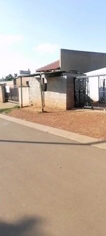 The video shows a still shot of a low-rise brick building with a red tile roof, as seen from a street. The building has a black metal gate on the right side. The ground in front of the building is reddish dirt. The street is paved and appears to be in a residential area. The sky is blue with scattered clouds. There are other buildings visible in the background. There are no people visible.