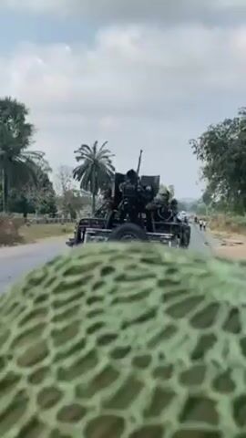 The video depicts a dark-colored pickup truck with a mounted gun driving down a paved road. The truck carries several soldiers. The video is taken from another vehicle, presumably following the truck. The camera occasionally focuses on the camouflage netting covering the helmet of the person filming. The environment consists of roadside vegetation, palm trees, and a few pedestrians. The sky is overcast. The truck with the soldiers continues driving down the road.