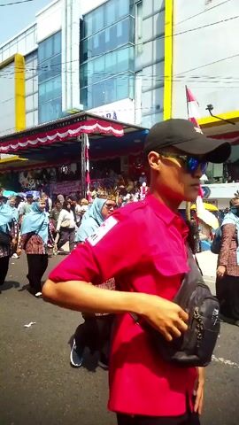 The video shows a street scene with several people. In the foreground, a young person wearing a red shirt, a black baseball cap, and sunglasses is walking from left to right. They have a black bag slung across their body. Behind them, a group of people, mostly women wearing headscarves and patterned clothing, are walking in the same direction. They appear to be part of a procession or parade. In the background, there is a building with a yellow and blue facade, and a stage decorated with red and white fabric. The overall impression is of an outdoor event or festival.
