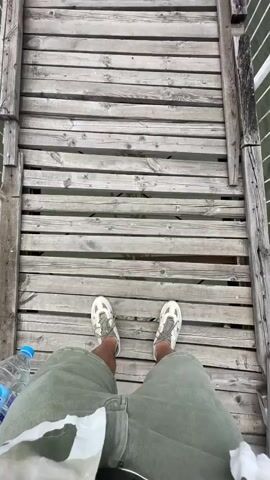 The video begins with a first-person view looking down at a person's feet on a wooden bridge, wearing olive green shorts and light-colored sneakers. A blue plastic water bottle is visible near the left foot. The camera then slowly pans up, revealing a long, narrow wooden suspension bridge stretching across a body of water. The bridge has white and blue metal railings on both sides and suspension cables overhead. In the distance, green, tree-covered hills or mountains are visible under an overcast sky, bordering the far side of the water.