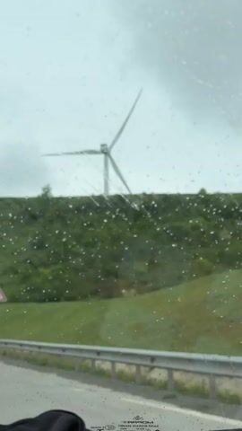 The video is shot from inside a car traveling on a road during a rainy day. Raindrops are visible on the windshield. Through the windshield, a wind turbine is visible on a green hill in the distance. There is a road sign with a left curve symbol on the roadside. The road has a gray metal guardrail. A black bag is visible on the car's dashboard.