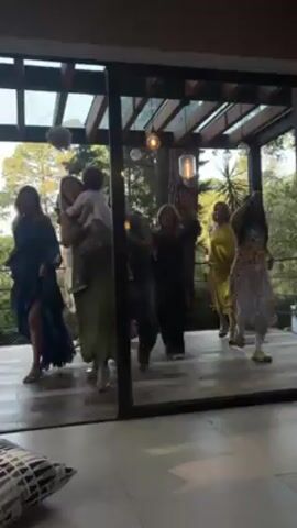 A group of about eight women and one child are dancing outside on a patio that is visible through a glass wall. The women are of various ages and sizes, and they are all wearing dresses. One of the women is holding a toddler. A young girl in a pink and white dress is in the foreground, moving in and out of the camera's view. The patio is made of wood, and there are some plants and trees visible in the background. The women appear to be having fun and enjoying themselves. The mood of the scene is joyful and festive.