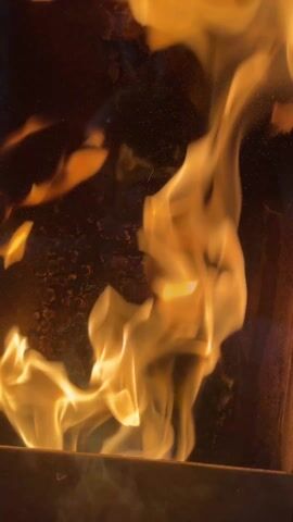 A close-up shot of a burning fireplace shows bright yellow flames dancing inside the firebox. The flames are quite large, reaching up towards the top of the frame. The interior of the firebox is dark, and the glass has a slightly frosted appearance.