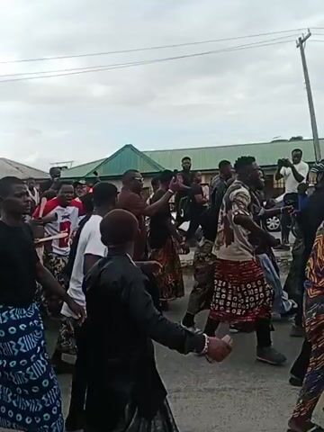 A group of people, most of them wearing black shirts and patterned skirts, are walking down a street. Some are dancing and raising their hands. There is a red car in the foreground, and buildings with green roofs in the background. The sky is overcast.