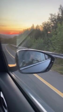 The video shows a view from inside a car driving on a road during sunset. The camera is positioned looking out the side window and then the windshield. In the side view, a side mirror reflects the interior of the car, showing a person. The road is paved with yellow and white lines. Trees and hills are visible on either side of the road, and the sky is filled with the colors of a sunset, with shades of orange, red, and yellow. The sun is low on the horizon. The video captures the movement of the car as it travels along the scenic route.