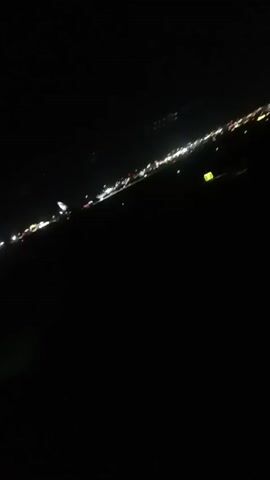 The view from an airplane window at night shows a strip of city lights in the distance. The lights are mostly white, with some red and green. The ground below is dark and indistinguishable. The camera is slightly angled. The scene is mostly still.