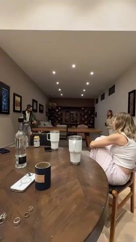 The video is a first-person perspective from a seated position at a round wooden table. In the foreground, on the table, are a glass bottle of clear liquid, two large white tumblers, a dark blue cylindrical object, and a smartphone. Several rings are also visible on the table's surface. In the mid-ground, a long wooden table, possibly a ping pong table, is set up. Two individuals are playing ping pong at the far end of this table. One person, wearing a green jacket and a cap, is standing and appears to be playing. The other person, wearing a light-colored top and dark pants, is also standing and facing the first person, seemingly engaged in the game. In the background, a woman is seated at the end of the long table, facing away from the camera and towards the ping pong game. She is wearing a white top and light-colored pants, and her legs are crossed. The environment appears to be an indoor recreational space or a living room with artwork on the walls, shelves filled with books, and recessed lighting in the ceiling. The overall scene depicts a casual gathering with a game of ping pong in progress.