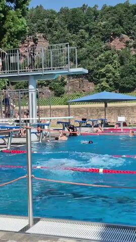 At 00:00, a person is seen jumping from a high diving board. At 00:01, the person is in mid-air, performing a dive. At 00:02, the person is entering the water, creating a splash. At 00:03, several people are swimming in the pool, with lane markers visible. At 00:04, more people are visible on the diving board and in the water, with trees and a rocky cliff face in the background.