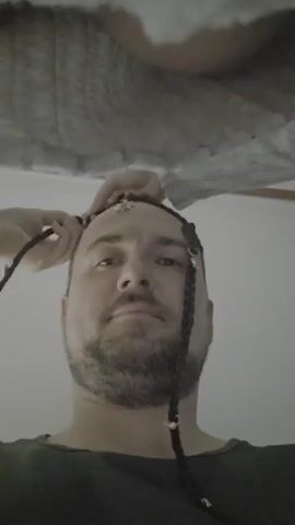 A low-angle, close-up shot shows a man's face as someone's hands braid his hair. The man has short brown hair and a beard, and he is wearing a dark green shirt. A few decorative beads are attached to the braids. The background is out of focus, but appears to be an indoor setting with a textured surface above.