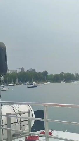 The video shows a view from a boat, looking out over a body of water. In the foreground, there is a railing with a white life raft. Beyond the railing is a calm body of water where several sailboats are anchored. In the background, there is a tree-lined shore with some buildings visible in the distance. The sky is overcast. The person speaking in the audio is pointing out a blue pole in the water.
